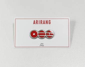 Puede incluir: Un pin rojo y plateado con la palabra "ARIRANG" impresa en rojo sobre una tarjeta blanca. El pin presenta tres formas circulares y rectangulares rojas y plateadas. Las palabras "yellow pinbadge" están impresas en letra pequeña.