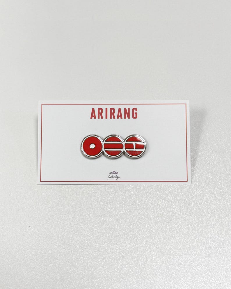 K&ouml;nnte beinhalten: Ein roter und silberner Anstecker mit dem Wort "ARIRANG" in Rot auf einer wei&szlig;en Karte gedruckt. Der Anstecker zeigt drei rote und silberne kreisf&ouml;rmige und rechteckige Formen. Die Worte "yellow pinbadge" sind in kleiner Schrift gedruckt.
