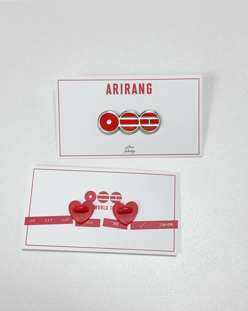 K&ouml;nnte beinhalten: Zwei wei&szlig;e Karten mit roten Akzenten. Die obere Karte zeigt das Wort "ARIRANG" &uuml;ber einer rot-wei&szlig;en Anstecknadel. Die untere Karte hat zwei herzf&ouml;rmige rote Anstecknadeln und die Worte "WORLD TOUR".
