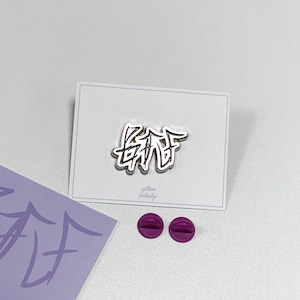 Puede incluir: Un pin plateado con la palabra "BAF" en una fuente estilizada, presentado en una tarjeta blanca. Dos cierres de pin morados están junto a la tarjeta. También es visible una pegatina morada con el mismo texto.