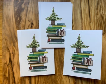 May include: Greeting cards featuring a watercolor illustration of a Christmas tree atop a stack of books. The tree is green with red berries, and the books are various shades of green, blue, and brown. A small gift box with a red bow is also included.