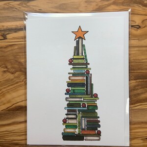以下が含まれることがあります： 積み重ねられた本で作られたクリスマスカード。さまざまな色の本で構成された木で、オレンジ色の星が飾られ、赤いオーナメントで飾られています。カードは透明なプラスチック製のスリーブに入っています。