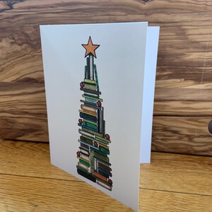 以下が含まれることがあります： 積み重ねられた本で作られたクリスマスツリーのデザインが特徴の白いグリーティングカード。木の上にはオレンジ色の星があり、赤いオーナメントで飾られています。カードは開いており、空白の内側が見えます。