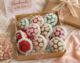 Könnte beinhalten: Eine Geschenkschachtel aus Pappe gefüllt mit mehreren gehäkelten Blumen-Schlüsselanhängern mit Büscheln aus rosa, roten und weißen Garnblüten an grünen Stielen. Jeder Anhänger hat einen Metallverschluss. Eine Karte zeigt Handmade with love und ein Etikett Especially for you.