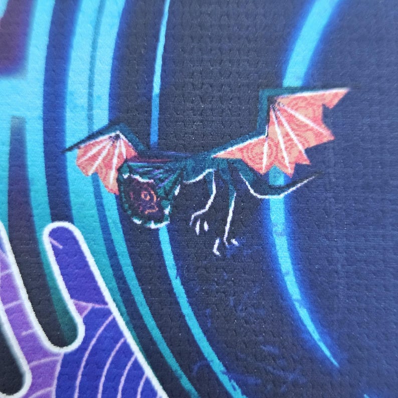 Puede incluir: Una superficie de tela azul oscuro con un gr&aacute;fico impreso de una peque&ntilde;a criatura con alas color coral y cuerpo oscuro. El fondo incluye l&iacute;neas y formas curvas abstractas en azul claro y morado que crean sensaci&oacute;n de movimiento y profundidad.