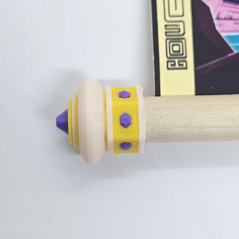 Puede incluir: Un remate de madera para pergamino con un dise&ntilde;o en capas, una punta morada, una base circular color crema y una banda cil&iacute;ndrica amarilla con peque&ntilde;os tachones hexagonales morados, unido a una varilla de madera de color claro.