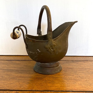 May include: A vintage copper coal scuttle with a handle and a ceramic knob. The scuttle has a worn, aged patina and sits on a round base.