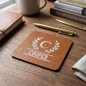 May include: A square brown faux leather coaster featuring a white embroidered design of a laurel wreath surrounding the letter C, with the name COOPER printed in white capital letters below the wreath on a horizontal line.