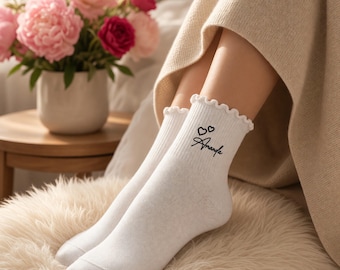 May include: White ankle socks featuring a ruffled top edge and black embroidered text that reads Amanda, accompanied by two small heart icons. The socks are made of a ribbed knit fabric and reach just above the ankle bone.