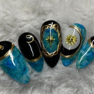 May include: Five long, almond-shaped nails with a blue and black marbled design. Each nail has a gold accent and a different celestial design: a star, a sun, and a crescent moon.