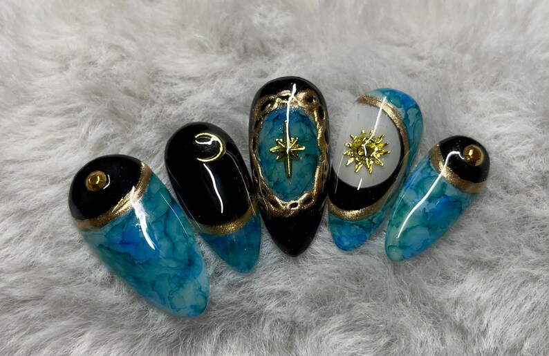 May include: Five long, almond-shaped nails with a blue and black marbled design. Each nail has a gold accent and a different celestial design: a star, a sun, and a crescent moon.