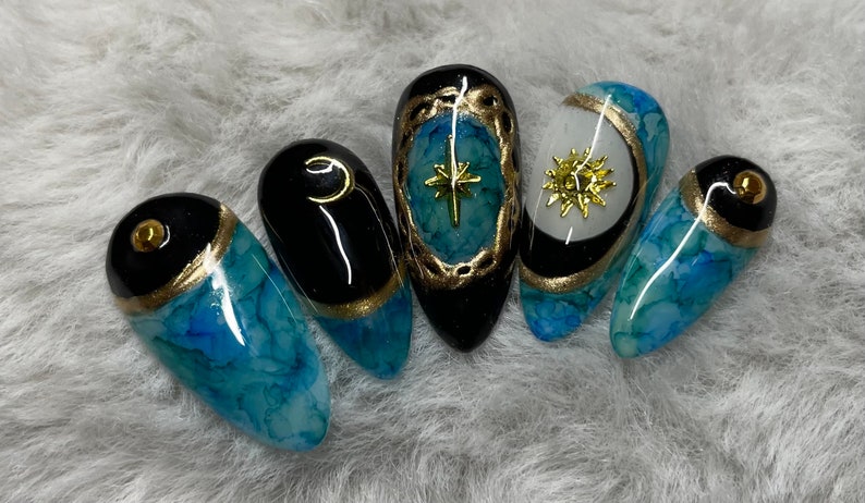 May include: Five blue and black nail tips with gold accents. Each nail tip features a different celestial design, including a crescent moon, a star, and a sun.