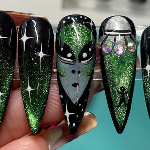 May include: Set of five long, pointed false nails. The nails feature a black and green glitter gradient design with white star accents. One nail has an alien face, another has a UFO with gems, and another has a stick figure.
