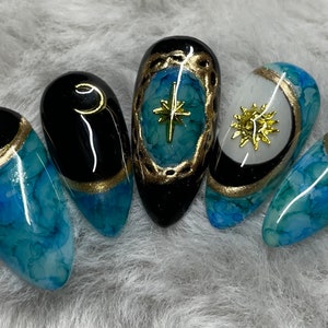 May include: Five blue and black almond-shaped nails with gold accents and celestial designs. The designs include a crescent moon, a star, and a sun.