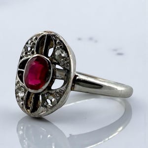 May include: An oval-shaped silver ring featuring a vibrant red ruby center stone. The ruby is surrounded by a decorative filigree design with small, clear accent stones. The ring's band is a simple, polished silver.