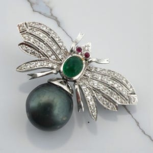 Puede incluir: Broche de abeja plateada con alas con diamantes, una piedra central verde ovalada y una gran perla gris oscura. Dos pequeñas piedras rojas están engastadas encima de la piedra verde.