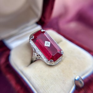 May include: A silver-coloured ring featuring a large, rectangular, deep red gemstone set in a filigree metal band. A small, square-shaped white diamond is embedded directly into the centre of the red stone, surrounded by intricate metalwork.