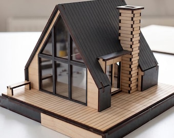 May include: A miniature A-frame house model constructed from light-colored wood with dark-toned accents. The structure features a dark roof, a tall chimney, large glass-paneled windows, and a wooden deck base. The model is assembled with visible joints.