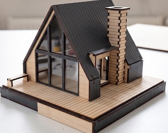 May include: A miniature A-frame house model constructed from light-colored wood with dark-toned accents. The structure features a dark roof, a tall chimney, large glass-paneled windows, and a wooden deck base. The model is assembled with visible joints.