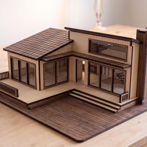 May include: A wooden architectural model of a modern house featuring a dark brown roof, light tan vertical siding, and large transparent windows. The structure includes a deck with a railing, a chimney, and a flat base with a wood grain pattern.