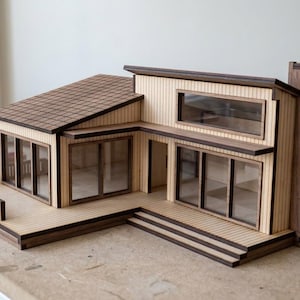 May include: A scale model of a modern house constructed from light-colored wood with dark wood trim. The structure features a sloped roof, a wrap-around deck with a railing, large rectangular windows, and a prominent chimney on the right side.