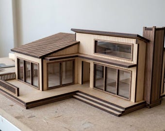May include: A scale model of a modern house constructed from light-colored wood with dark wood trim. The structure features a sloped roof, a wrap-around deck with a railing, large rectangular windows, and a prominent chimney on the right side.