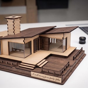 May include: A wooden architectural model of a house featuring a tall chimney, a slanted roof, and a deck. The base includes an engraved plaque with the text, A small memory for our family's father. The structure is made of light and dark wood panels.