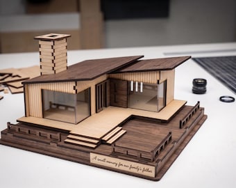 May include: A wooden architectural model of a house featuring a tall chimney, a slanted roof, and a deck. The base includes an engraved plaque with the text, A small memory for our family's father. The structure is made of light and dark wood panels.