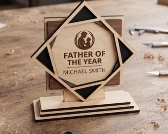 May include: A layered wooden award plaque featuring a geometric star design with a light wood center and dark wood accents. The center displays a circular icon of a parent and child with the text FATHER OF THE YEAR and the name MICHAEL SMITH.
