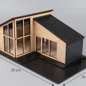 Pode incluir: Uma casa modelo de madeira com telhado preto e revestimento de madeira clara, apresentando grandes janelas e uma base preta. O modelo mede 25 cm de comprimento, 16 cm de largura e 12 cm de altura, conforme indicado pelas linhas na imagem.