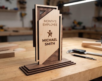 May include: A layered wooden award plaque featuring dark walnut and light maple wood. The front panel is engraved with the text MONTH'S EMPLOYEE, a star icon, and the name MICHAEL SMITH. The base includes the printed dimensions 180 x 65 x 195 mm.