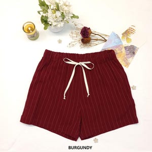 May include: Burgundy-colored shorts with vertical white pinstripes. The shorts have an elastic waistband with a white drawstring. The word "BURGUNDY" is printed at the bottom of the image. The shorts are displayed on a white surface.