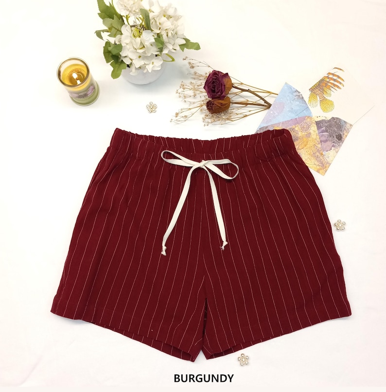 May include: Burgundy-colored shorts with vertical white pinstripes. The shorts have an elastic waistband with a white drawstring. The word "BURGUNDY" is printed at the bottom of the image. The shorts are displayed on a white surface.