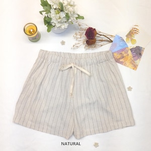 May include: Cream-colored shorts with vertical black stripes and a drawstring waist. The shorts are labeled "NATURAL" at the bottom. The image also includes a candle, flowers, and decorative elements.