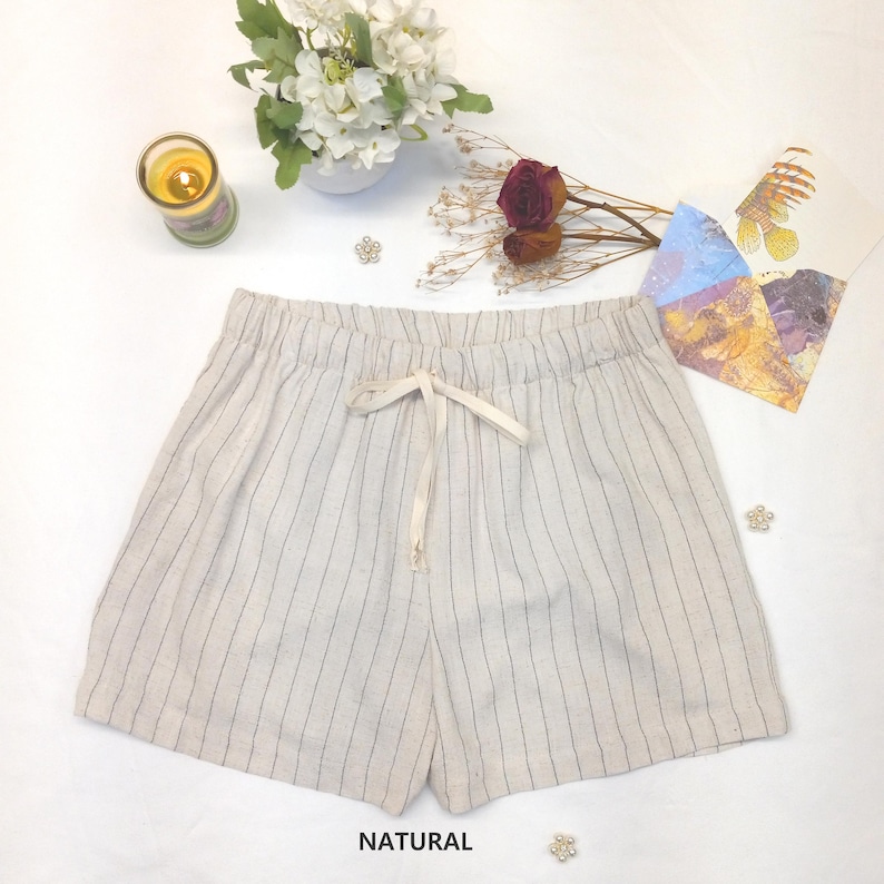 May include: Cream-colored shorts with vertical black stripes and a drawstring waist. The shorts are labeled "NATURAL" at the bottom. The image also includes a candle, flowers, and decorative elements.