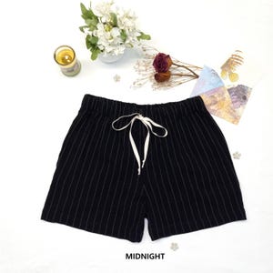 May include: Black shorts with vertical white pinstripes and a white drawstring. The shorts are labeled "MIDNIGHT" in white text. The shorts are on a white surface with decorative flowers, a candle, and other small items.