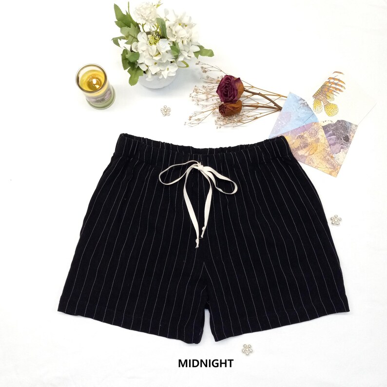 May include: Black shorts with vertical white pinstripes and a white drawstring. The shorts are labeled "MIDNIGHT" in white text. The shorts are on a white surface with decorative flowers, a candle, and other small items.