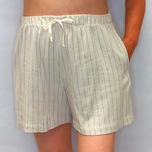 May include: Cream-colored shorts with vertical black pinstripes. The shorts have an elastic waistband with a drawstring and two side pockets. The fabric appears to be linen or a similar lightweight material, suitable for warm weather.