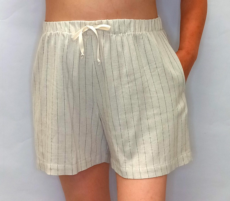 May include: Cream-colored shorts with vertical black pinstripes. The shorts have an elastic waistband with a drawstring and two side pockets. The fabric appears to be linen or a similar lightweight material, suitable for warm weather.