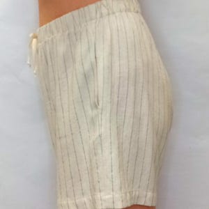 May include: Cream-colored shorts with vertical gray stripes. The shorts have an elastic waistband with a drawstring and a single pocket. The fabric appears to be linen or a similar lightweight material, suitable for warm weather.