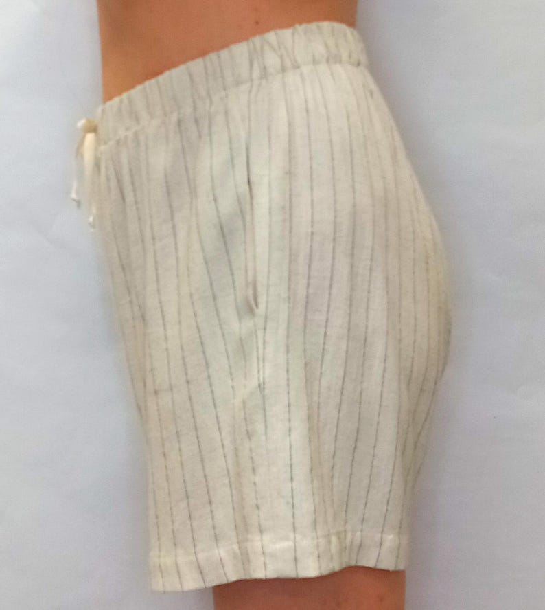 May include: Cream-colored shorts with vertical gray stripes. The shorts have an elastic waistband with a drawstring and a single pocket. The fabric appears to be linen or a similar lightweight material, suitable for warm weather.