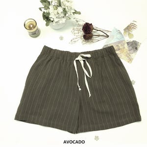 May include: Avocado-colored shorts with vertical white pinstripes and a white drawstring. The shorts have an elastic waistband. The word "AVOCADO" is printed at the bottom of the image. The shorts are displayed on a white surface.