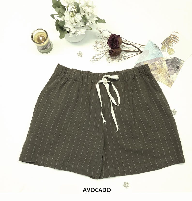 May include: Avocado-colored shorts with vertical white pinstripes and a white drawstring. The shorts have an elastic waistband. The word "AVOCADO" is printed at the bottom of the image. The shorts are displayed on a white surface.