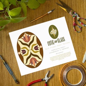 May include: A white paper sheet featuring a Folk Flower Panel Pattern by Cutie Pie Glass, displaying an oval design with brown, cream, and pink floral motifs. The text includes difficulty level, 68 pieces, and copyright information.