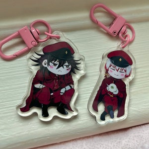May include: Two acrylic keychains featuring anime-style characters in maroon uniforms and hats. The keychains have pink clasps. One character is squatting, and the other is holding an object. The background is a light-colored surface.