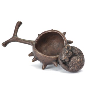 Op de afbeelding: Een bronzen sieradendoosje in de vorm van een kastanje, met een slapende muisfiguur als deksel. De doos heeft een getextureerd oppervlak en een takvormige handgreep. De muis is opgerold.