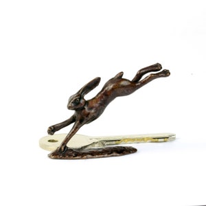 May include: A bronze sculpture of a hare leaping over a key. The hare is in mid-air, with its legs extended and its ears perked up. The sculpture is detailed and realistic, and the hare's fur is textured.