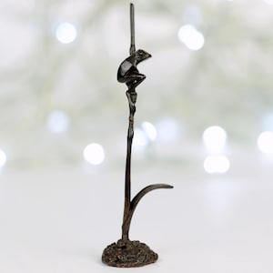 Könnte beinhalten: Eine kleine Bronzeskulptur eines Frosches, der auf einem hohen, dünnen Ast sitzt. Der Frosch ist nach rechts gewandt und hat eine detaillierte, realistische Darstellung. Die Skulptur steht auf einem kleinen Sockel.