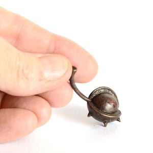 May include: A small, bronze, spiky, chestnut-shaped object with a curved handle. The object is being held in a hand.