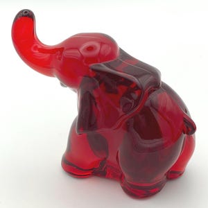 Puede incluir: Una figura de elefante de cristal rojo con la trompa levantada. La figura es de color rojo sólido y brillante con vetas rojas más oscuras. El elefante está sentado sobre sus patas traseras, con las orejas y la trompa claramente visibles.